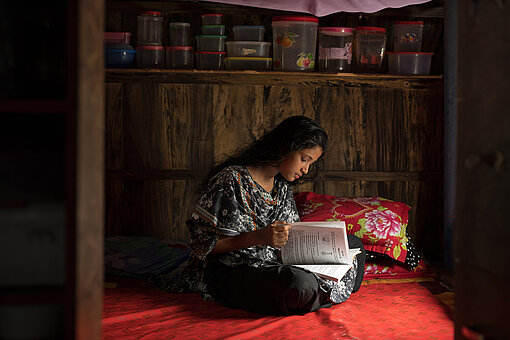 Ein jugendliches Mädchen aus Bangladesch sitzt auf einer roten Matratze in einem Haus und liest ein Buch