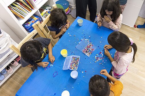Fünf Kinder sitzen an einem Tisch und ziehen Perlen auf Schnüre