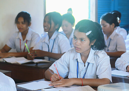 Schülerinnen in weißen Hemden sitzen im Klassenraum und schreiben in ihre Hefte, ein Mädchen ist im Fokus