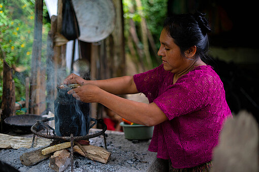 Eine Frau mit buntem Oberteil kocht etwas über einem kleinen Holzfeuer