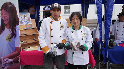 Ein junger Mann und eine junge Frau, beide gekleidet in professionelle weiße Kochjacken, stehen nebeneinander und halten jeweils Lebensmittelverpackungen in den Händen. Sie wirken stolz und selbstbewusst, als würden sie ihre Produkte auf einer Messe oder Ausstellung präsentieren