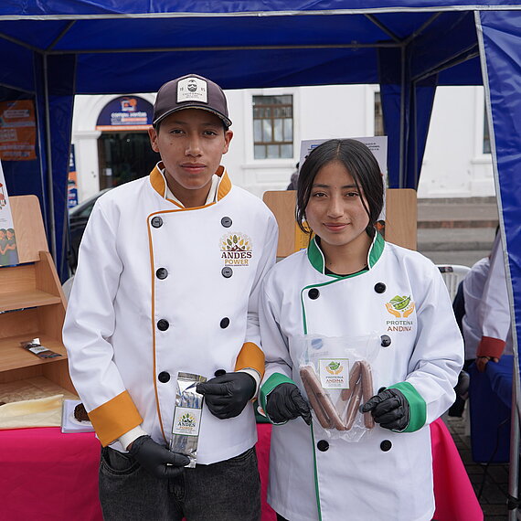 Ein junger Mann und eine junge Frau, beide gekleidet in professionelle weiße Kochjacken, stehen nebeneinander und halten jeweils Lebensmittelverpackungen in den Händen. Sie wirken stolz und selbstbewusst, als würden sie ihre Produkte auf einer Messe oder Ausstellung präsentieren