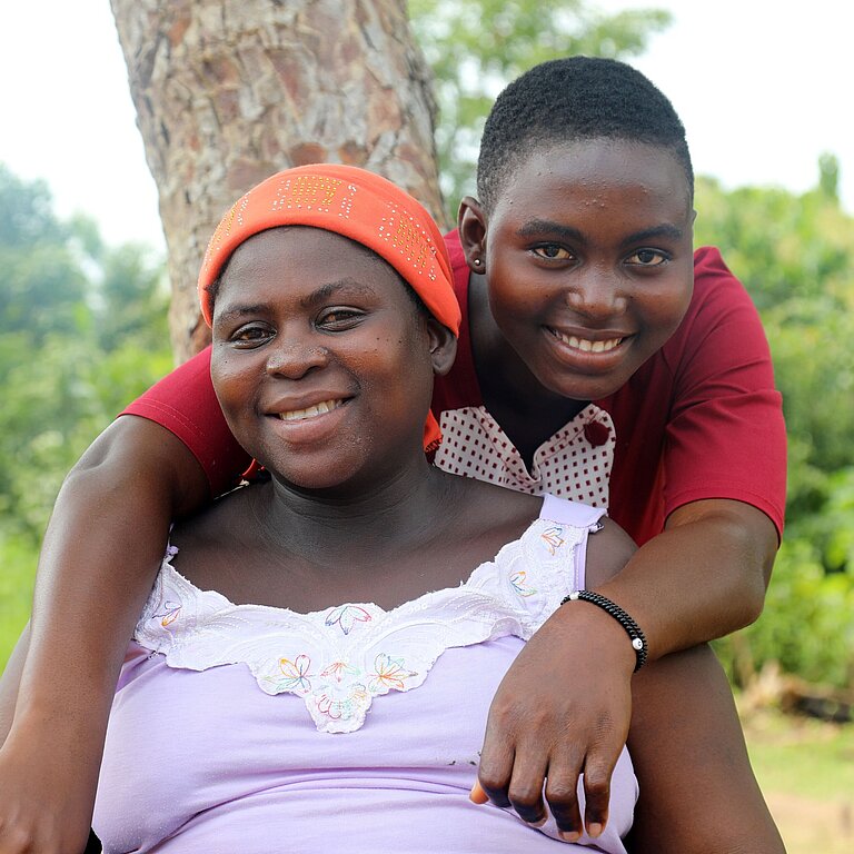 Eine junge Frau mit rotem T-Shirt umarmt eine ältere Frau, die im Garten auf einer Bank sitzt und ein weißes Shirt trägt, und beide lächeln