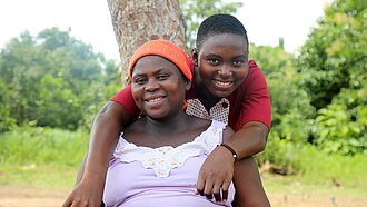 Eine junge Frau mit rotem T-Shirt umarmt eine ältere Frau, die im Garten auf einer Bank sitzt und ein weißes Shirt trägt, und beide lächeln