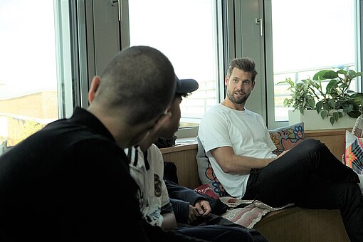 Zwei Schüler sitzen auf einer Bank und schauen Jan Möller an. Dieser sitzt ebenfalls auf der Bank und spricht mit den Schülern.