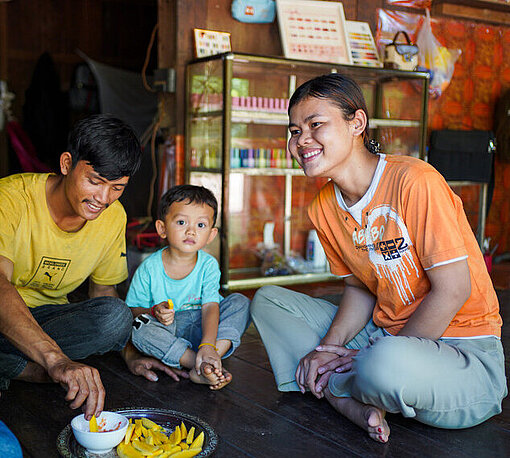 Ein Mann, eine Frau und ein junges Kind sitzen auf dem Boden eines Schönheitssalons und essen Früchte aus einer großen Schale.