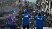 Drei Personen mit blauen Kappen und blauen Westen mit dem Logo von Plan International sowie eine weitere Person stehen vor einer zerstörten, schlammigen Straße zwischen beschädigten Häusern. Im Hintergrund sind weitere Menschen und Berge zu sehen.