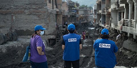 Drei Personen mit blauen Kappen und blauen Westen mit dem Logo von Plan International sowie eine weitere Person stehen vor einer zerstörten, schlammigen Straße zwischen beschädigten Häusern. Im Hintergrund sind weitere Menschen und Berge zu sehen.