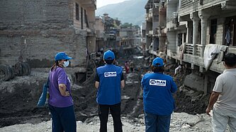 Drei Personen mit blauen Kappen und blauen Westen mit dem Logo von Plan International sowie eine weitere Person stehen vor einer zerstörten, schlammigen Straße zwischen beschädigten Häusern. Im Hintergrund sind weitere Menschen und Berge zu sehen.