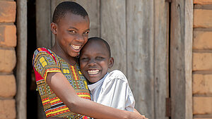 Zwei lachende Kinder stehen eng umarmt vor einer einfachen Ziegelwand und einer Holztür und schauen fröhlich in die Kamera.