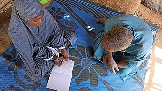 Ein Mädchen sitzt auf einer Matte und hält ein Notizbuch, in dem sie schreibt. Ein Mann sitzt bei ihr.