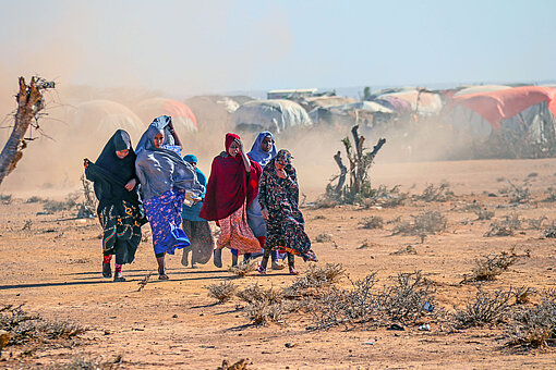 Wegen anhaltender Dürre mussten viele Menschen in Somalia ihre Heimat verlassen, auf der Sucher nach Wasser, Nahrung und Arbeit. Eine Gruppe Mädchen in bunten Gewändern läuft durch eine staubige Wüstenlandschaft. Im Hintergrund ist eine Zelt-Siedlung zu erkennen.