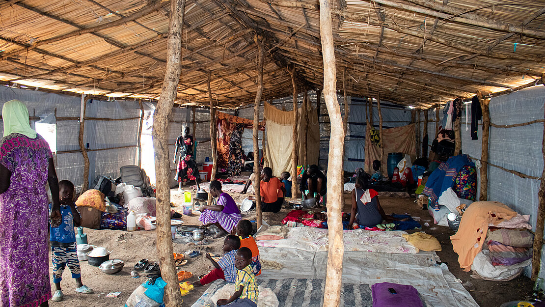 Ein Shelter von innen Im inneren eines großem Zeltes sitzen mehrere Menschen auf dem Boden auf Decken, eine Frau und ein Kind stehen links im Bild, an einigen Stöcken sind Tücher als Sichtschutz aufgehängt