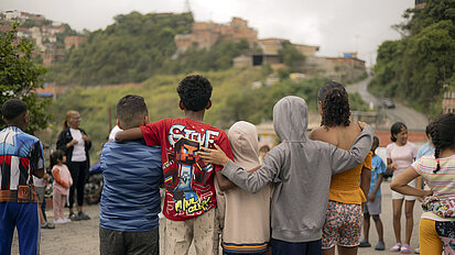 ine Gruppe von Kindern steht Arm in Arm mit dem Rücken zur Kamera auf einer Straße. Im Hintergrund sind weitere Kinder und Erwachsene sowie eine hügelige Vorstadtszene zu sehen.