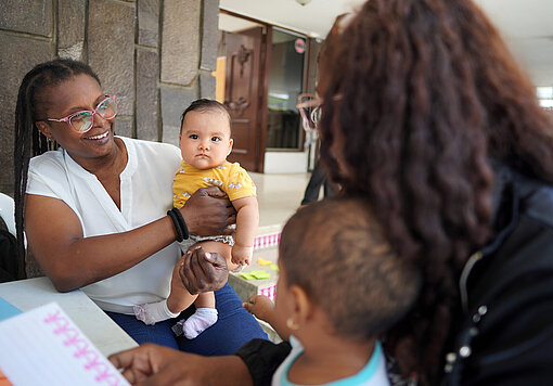 Eine Frau hält ein kleines Kind im Arm, während eine zweite Person mit einem weiteren Kind am Tisch sitzt. Im Hintergrund ist eine Tür und ein offener Raum sichtbar.