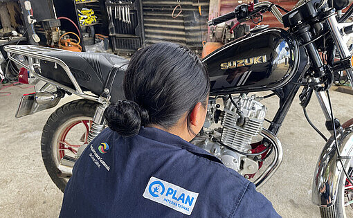 Eine junge Frau in Blaumann hockt vor einem schwarzen Motorrad