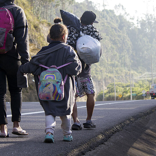 Eine Frau und zwei Kinder laufen am Rande einer Schnellstraße. Sie tragen Rucksäcke.