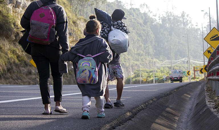 Eine Frau und zwei Kinder laufen am Rande einer Schnellstraße. Sie tragen Rucksäcke.