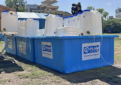Mehrere große und kleine Kanister mit einem blauen Plan-Logo sind eine provisorische Trinkwasserstation