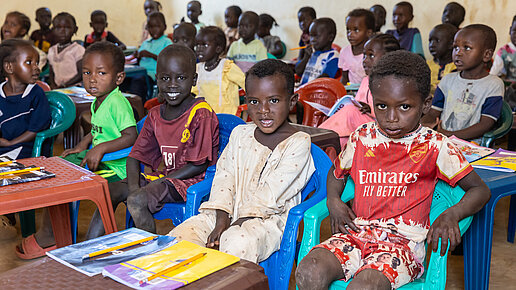 Guppe von Kindern sitzt in einem Klassenzimmer. Im Vordergrund sind vier Jungs zu sehnen die vor ihren Schulheften sitzen.