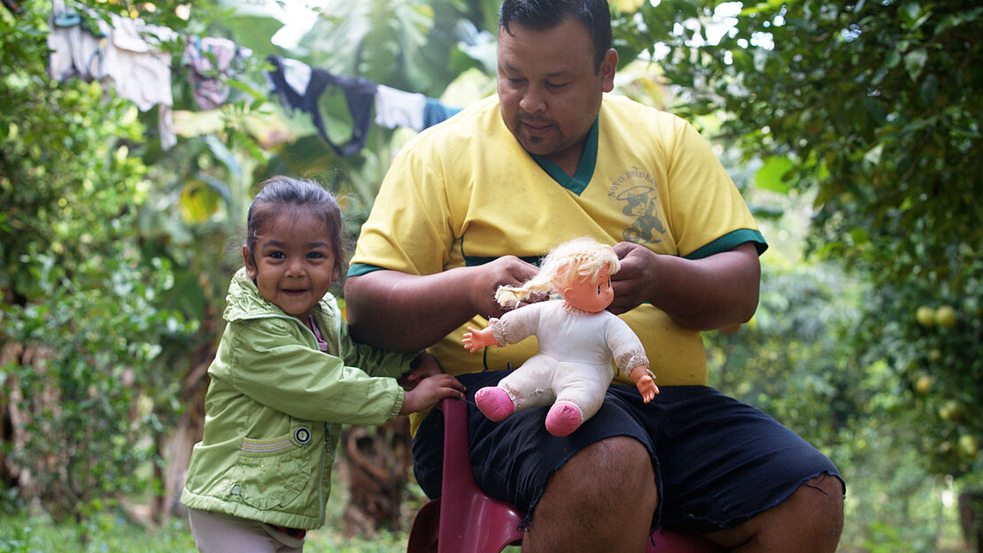 Christian sitzt im Garten und spielt mit seiner Tochter und ihrer Puppe.