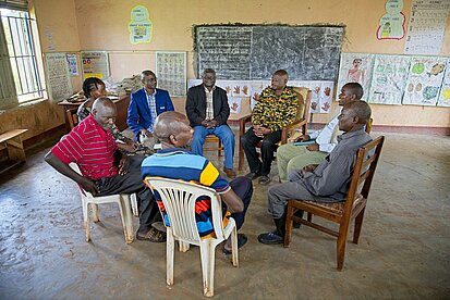 Eine Gruppe erwachsener Männer sitzt im Kreis in einem Klassenraum, einige tragen Anzüge, andere Hemden oder bunte Shirts. Sie befinden sich in einer Gesprächssituation.