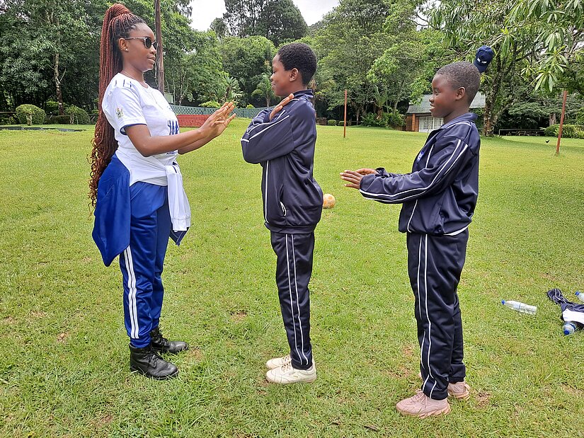 Eine Frau in Sportkleidung interagiert mit zwei Kindern in Trainingsanzügen, die sich auf einem grünen Feld gegenüberstehen. Die Szene scheint Teil einer Sport- oder Teamübung zu sein.