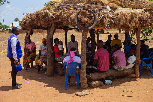 Mehrere Menschen sitzen unter einem kleinen Pavillion aus Baumstämmen und Stroh, davor steht ein Mitarbeiter von Plan International in blauer Weste