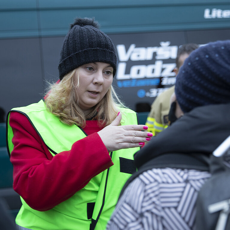 Monica steht in gelber Warnweste vor einem Bus und spricht mit Menschen.