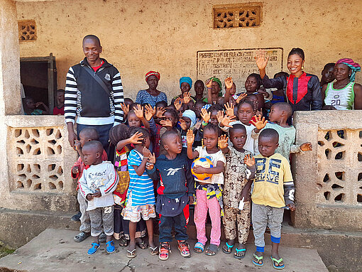 Tamba Michel (links) mit Kindern einer von Plan International unterstützten Vorschule Eine Gruppe Kinder steht vor einem Schulgebäude