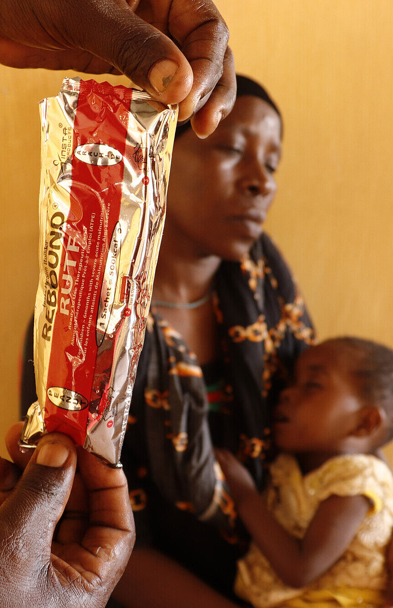 Im Vordergrund wird eine Plastiktüte in die Kamera gehalten, in der die Ernährungspaste ist. Im Hintergrund sitzt eine Mutter mit einem kleinen Kind im Schoß.