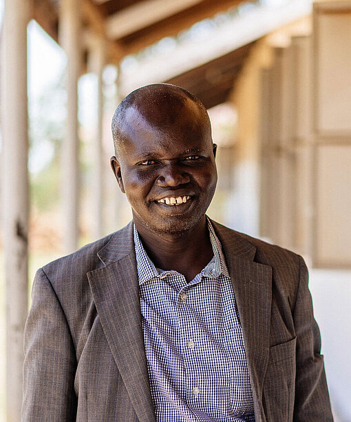 Lehrer Matthew Amanzuru hat die besonderen Herausforderungen von Mädchen im Blick. Ein Lehrer in Hemd und Sakko steht vor dem Schulgebäude und lächelt in die Kamera.