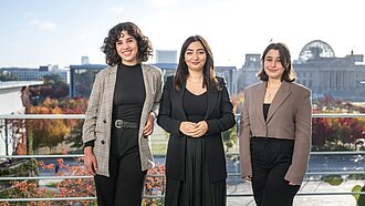 Zwei junge Frauen stehen an der Seite der Integrationsstaatsministerin auf der Terasse der Kanzleramts.