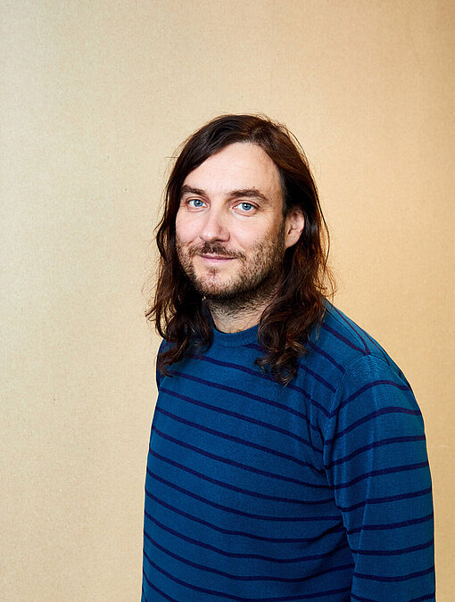 Portraitfoto von einem Mann mit braunen, langen Haaren, der freundlich in die Kamera schaut. Er trägt einen blauen Pullover und steht vor einem neutralen Hintergrund.