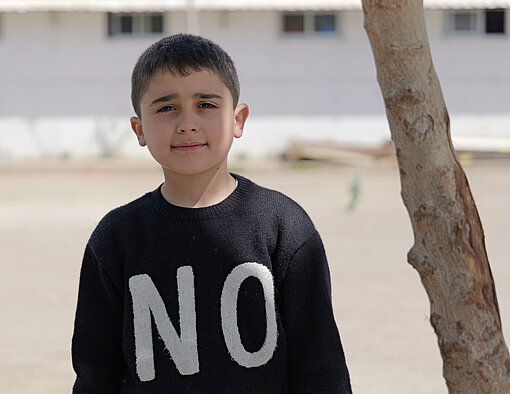 Ein Kind steht draußen vor einem hellen Gebäude, trägt einen schwarzen Pullover mit weißen Buchstaben und blickt in die Kamera. Ein Baum ist am rechten Bildrand zu sehen.