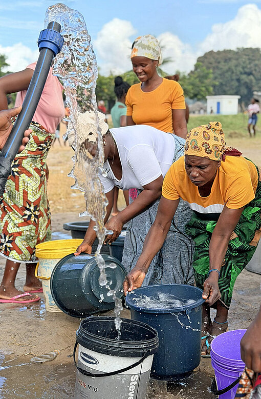 Frauen mit Plastikeimern in den Händen drängen um einen Schlauch, aus dem Wasser sprudelt