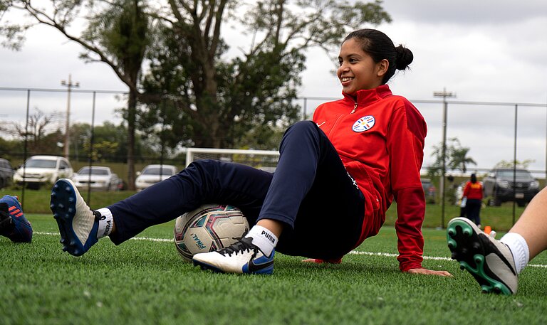 Mädchen in rotem Trainingsanzug sitzt auf dem Fußballrasen und wärmt sich mit einem Fußball auf