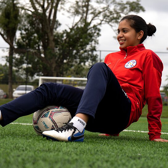 Mädchen in rotem Trainingsanzug sitzt auf dem Fußballrasen und wärmt sich mit einem Fußball auf