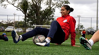 Mädchen in rotem Trainingsanzug sitzt auf dem Fußballrasen und wärmt sich mit einem Fußball auf