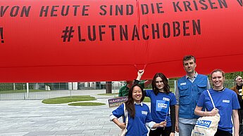 Vor einem roten Protestbanner stehen vier Mitarbeiter:innen von Plan International