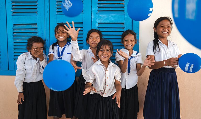 Fröhliche Schulkinder in Schuluniformen stehen vor einer blauen Wand und spielen lachend mit blauen Luftballons.