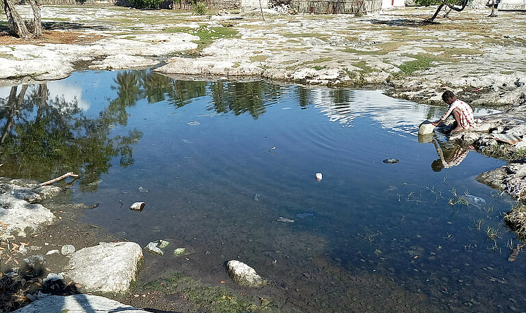 Ein Tümpel, an dem ein Mädchen sitzt und Wasser schöpft