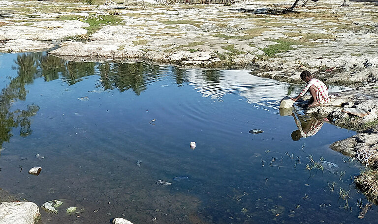 Ein Tümpel, an dem ein Mädchen sitzt und Wasser schöpft