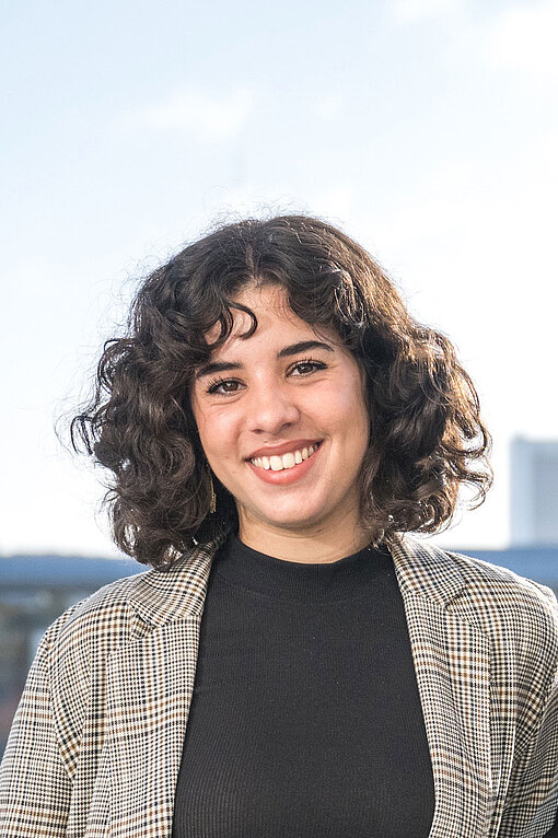 Lale berichtet über die feministische Revolution im Iran - und die Gefahr, in die sich Aktivistinnen vor Ort begeben. Portrait von Lale - sie trägt ein schwarzes Shirt und einen beigefarbenen karierten Blazer und lächelt in die Kamera.