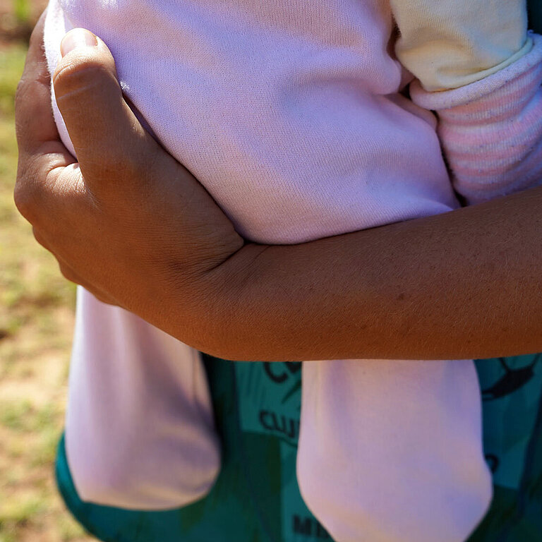 Eine Nahaufnahme von einem gebräunten Arm, der ein Baby in einem rosa Strampler hält. 