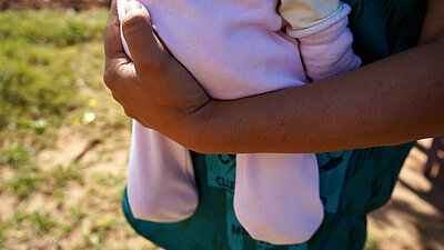 Eine Nahaufnahme von einem gebräunten Arm, der ein Baby in einem rosa Strampler hält. 