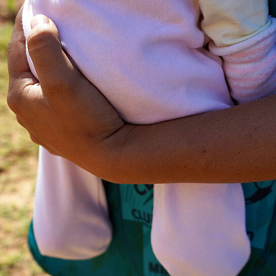 Eine Nahaufnahme von einem gebräunten Arm, der ein Baby in einem rosa Strampler hält. 