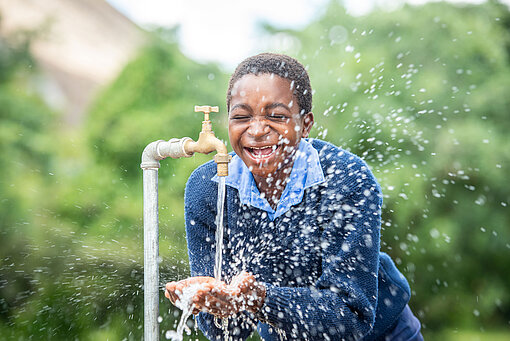Die Patenschaftsbeiträge fließen in Projekte in der Gemeinde, in der das Patenkind wohnt. Ein Mädchen in Schuluniform hält seine Hände unter einen laufenden Wasserhahn. Das Wasser spritzt in alle Richtungen und das Mädchen lacht.
