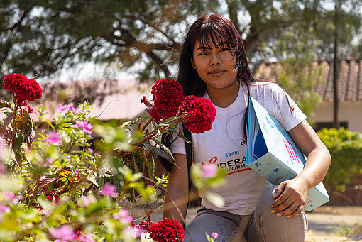 Ein Mädchen kniet neben einem Rosenbusch. Sie hat einen Aktenordner unter dem Arm.