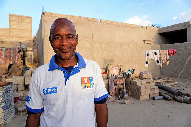 Ein Mann mit Plan T-Shirt steht in Timbuktu in einer Wohngegend zwischen Häusern. Hinter ihm hängen Anziehsachen an einer Wäscheleine.